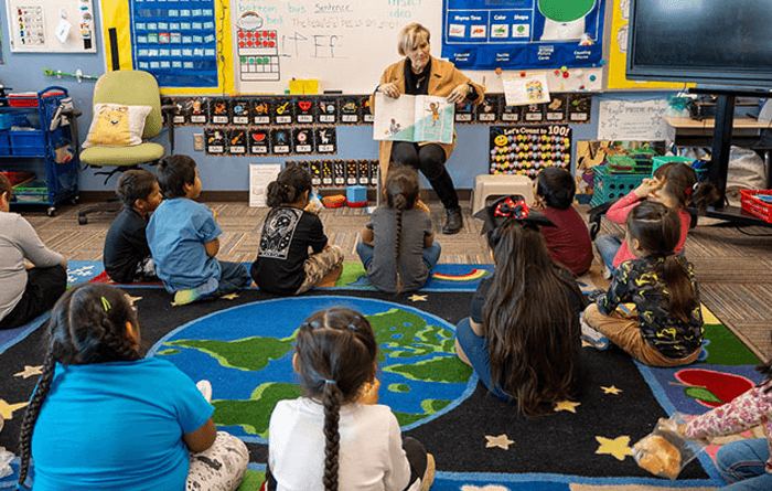 Teacher reading a book to a students sitting on the floor in a classroom.