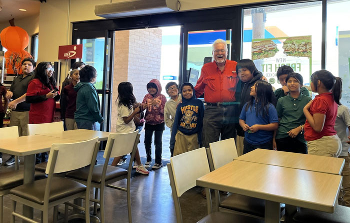An older man in a red shirt stands smiling with a group of elementary school–aged children inside a fast-food restaurant.