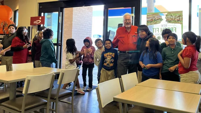 An older man in a red shirt stands smiling with a group of elementary school–aged children inside a fast-food restaurant.