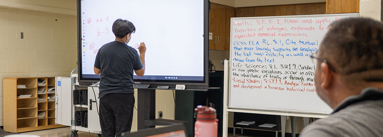 Male student writing on a digital board while male teacher watches the math problem. 