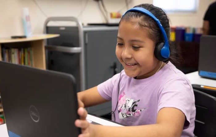 A student wearing headphones smiles while focused on laptop screen, engaged in an online activity.