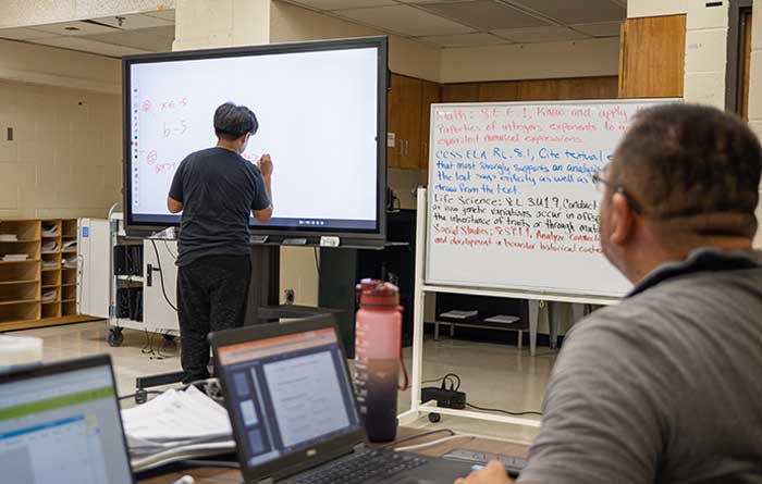 Student writing on an interactive whiteboard in a classroom while a teacher observes.