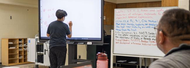 Male student writing on a digital board while male teacher watches the math problem. 