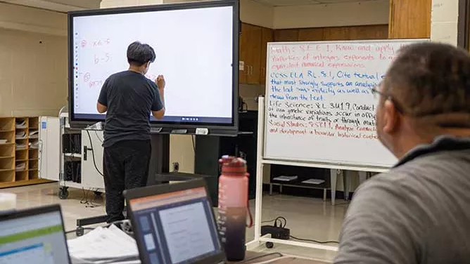 Student writing on an interactive whiteboard in a classroom while a teacher observes.